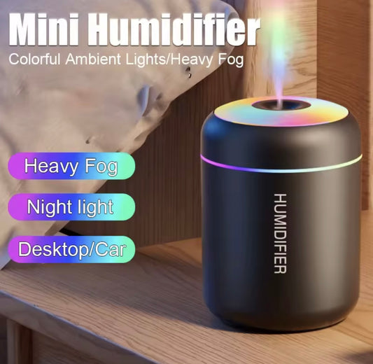 A black mini humidifier with a cylindrical body and a misting top, shown on a wooden surface with a blurred bedroom background. The image includes text overlays reading 'Mini Humidifier', 'Colorful Ambient Lights/Heavy Fog', 'Heavy Fog', 'Night light', and 'Desktop/Car'.